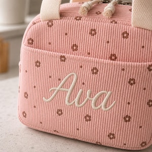 May include: A pink corduroy lunch bag featuring a repeating pattern of small brown flowers. The front pocket is embroidered with the name Ava in cream-colored script. The bag includes cream-colored fabric handles and a zipper with two knotted pulls.