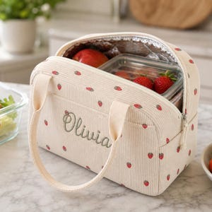 May include: A cream-colored corduroy lunch bag featuring a repeating strawberry pattern and the name Olivia embroidered in green script. The bag has two fabric handles, a zippered top closure, and a silver insulated interior containing food.