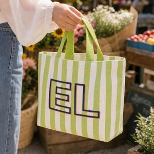 May include: A rectangular tote bag featuring vertical lime green and white stripes with two matching lime green fabric handles. The front of the bag displays the large capital letters EL in navy blue with a thin orange outline centered on the stripes.