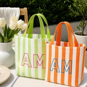 May include: Two canvas tote bags with vertical stripes and top handles. The left bag is light green and white with pink letters reading AM AM. The right bag is orange and white with blue letters reading AM AM. Both bags feature a rectangular shape.