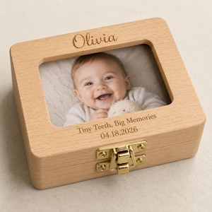 Könnte beinhalten: Eine helle Holz-Erinnerungsbox mit einem rechteckigen Sichtfenster, das ein lächelndes Baby zeigt. Der Deckel ist mit dem Namen Olivia graviert, und auf der Basis steht der Text Tiny Teeth, Big Memories sowie das Datum 04.18.2026. Ein goldener Metallverschluss sichert die Vorderseite.