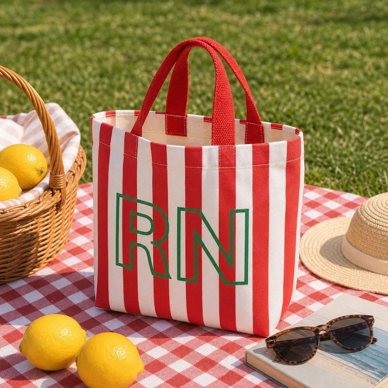 May include: A canvas tote bag featuring vertical red and white stripes with two red fabric handles. The front of the bag displays the letters RN in a bold, dark green outline font. The bag has a rectangular shape and a flat base.