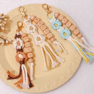 Puede incluir: Tres llaveros hechos a mano con cuentas de madera que deletrean nombres e iniciales. Cada llavero presenta un diseño de flores de macramé en varios colores, incluyendo marrón, crema y azul. Los cierres dorados están unidos a la parte superior de cada llavero.