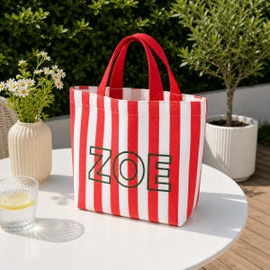 May include: A rectangular tote bag featuring vertical red and white stripes with two red fabric handles. The front of the bag displays the name ZOE in large, bold, dark green outlined capital letters centered on the striped pattern.