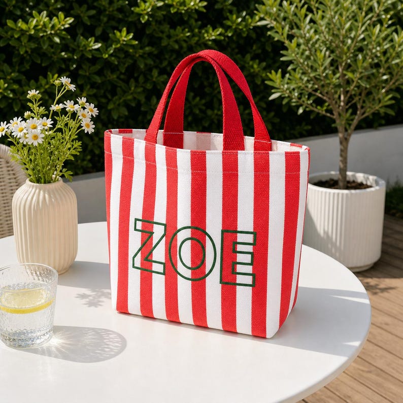 May include: A rectangular tote bag featuring vertical red and white stripes with two red fabric handles. The front of the bag displays the name ZOE in large, bold, dark green outlined capital letters centered on the striped pattern.