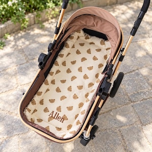 May include: A baby stroller featuring a cream-colored mattress pad with a repeating brown teddy bear pattern. The name Allen is embroidered in brown script on the mattress. The stroller has a brown canopy and a gold-toned metal frame.