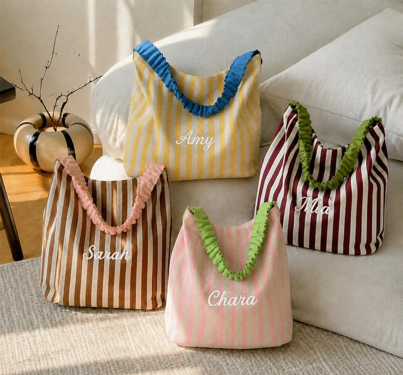 May include: Four fabric tote bags with vertical stripes and ruffled handles. The yellow bag features the name Amy, the brown bag features Sarah, the pink bag features Chara, and the maroon bag features Mia. Each bag has a soft, unstructured shape.