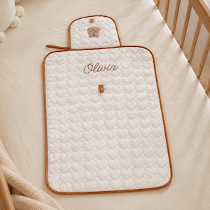 May include: A rectangular white quilted changing mat featuring an embossed heart pattern and brown trim. The name Olivia is embroidered in brown script in the centre. A small flap at the top displays a brown bear icon and a single brown button.