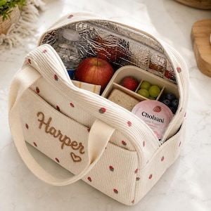May include: A cream-colored corduroy lunch bag with a small strawberry print, featuring the name Harper embroidered in brown thread above a small heart. The interior is lined with silver thermal insulation and contains a water bottle and food.