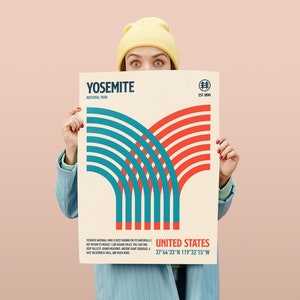 May include: A poster featuring a graphic design with red and teal lines, with the text "YOSEMITE NATIONAL PARK" and "UNITED STATES". The poster has a cream background and is held by a person wearing a yellow beanie and blue blazer.