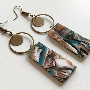 Può includere: Un paio di orecchini pendenti con un ciondolo rettangolare marmoreo marrone e bianco e un anello dorato con un piccolo ciondolo rotondo.