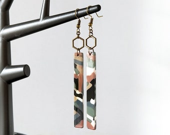 May include: A pair of long, rectangular earrings with a geometric pattern in shades of pink, green, black, and white. The earrings are made of polymer clay and have a brass hook and hexagonal charm.