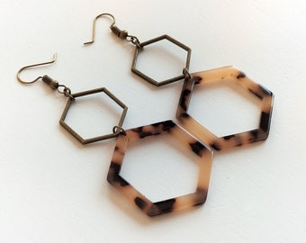 Könnte beinhalten: Ein Paar sechseckige Ohrringe mit einem Schildpattmuster. Die Ohrringe sind aus hellbraunem und schwarzem Material gefertigt und haben einen bronzefarbenen Metallhaken.
