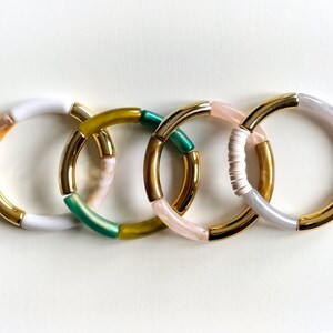 Può includere: Quattro braccialetti colorati con accenti dorati. I braccialetti sono fatti di perline bianche, verdi, gialle e rosa.