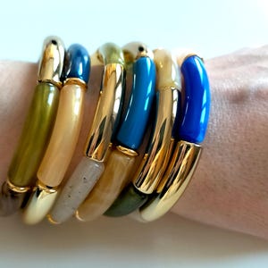 Könnte beinhalten: Ein Set aus bunten Armreifen mit goldenen Akzenten. Die Armreifen sind auf dem Handgelenk einer Person gestapelt und in verschiedenen Farben gehalten, darunter Blau, Grün und Beige.