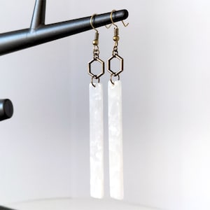 Op de afbeelding: Witte rechthoekige oorbellen met een zeshoekige hanger die van boven hangt. De oorbellen zijn gemaakt van een wit materiaal met een marmer effect.