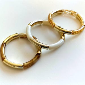 Peut inclure: Trois bracelets dorés et blancs. L'un des bracelets présente un motif or et marron, un autre est blanc avec des accents dorés, et le troisième est doré avec des paillettes. Les bracelets sont sur une surface blanche.