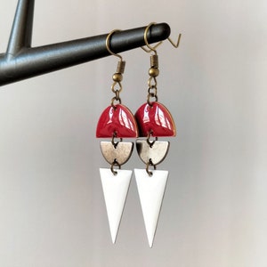 Può includere: Un paio di orecchini geometrici con uno schema di colori rosso, bianco e oro. Gli orecchini presentano una forma a semicerchio, una forma a mezzaluna e una forma triangolare.
