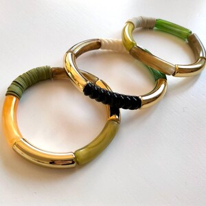 Può includere: Tre braccialetti dorati con diversi accenti in plastica e tessuto colorati. I braccialetti sono impilati uno sopra l'altro. Il braccialetto superiore è verde, bianco e nero. Il braccialetto intermedio è nero e verde. Il braccialetto inferiore è arancione, verde e oro.