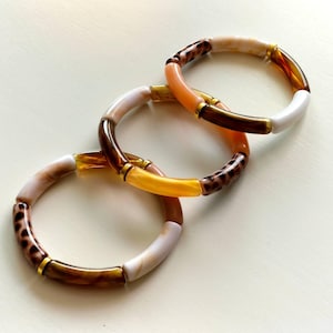 Puede incluir: Tres pulseras tubulares coloridas. Cada pulsera presenta una mezcla de colores, incluyendo blanco, marrón, naranja y amarillo. Las pulseras tienen detalles dorados y un estampado de leopardo. Las pulseras están hechas de un material brillante.
