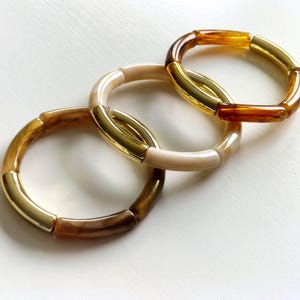 Könnte beinhalten: Drei verbundene Armbänder. Jedes Armband besteht aus Segmenten in Braun-, Creme- und Goldtönen. Die Armbänder sind auf einer weißen Oberfläche angeordnet und bilden eine ansprechende Schmuckpräsentation.