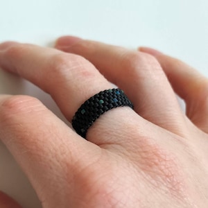 Può includere: Un anello nero di perline con piccoli accenti turchese e blu. L'anello è realizzato con piccole perline fittamente intrecciate, creando una superficie strutturata. L'anello è indossato su un dito, mettendo in mostra l'intricato lavoro di perline e le variazioni di colore.