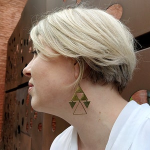 Könnte beinhalten: Eine Frau trägt ein weißes Hemd mit großen goldenen Dreiecksohrringen. Die Ohrringe bestehen aus mehreren übereinanderliegenden Dreiecken.