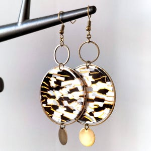 Peut inclure: Une paire de boucles d'oreilles en ton or avec un motif rond noir et blanc. Les boucles d'oreilles ont un petit disque doré qui pend en bas.
