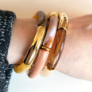 Könnte beinhalten: Drei goldene Armreifen mit verschiedenen farbigen Harzeinsätzen. Die Armreifen sind auf dem Handgelenk einer Person gestapelt. Die Harzeinsätze sind braun, beige und weiß.