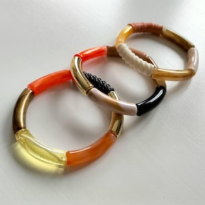 Puede incluir: Tres pulseras coloridas con una mezcla de cuentas naranjas, amarillas, negras y blancas. Las pulseras están hechas de plástico y tienen un acento de metal dorado.