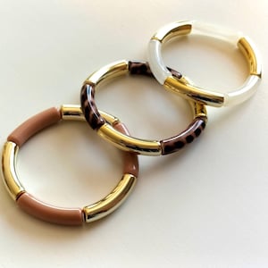 May include: Three stacked bracelets with gold-tone accents. One bracelet is light pink, another has a brown leopard print, and the third is white. The bracelets are made of a smooth, glossy material.