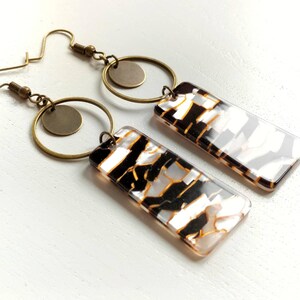 Könnte beinhalten: Ein Paar goldene Creolen mit einem abstrakten Design in Schwarz und Weiß. Die Creolen haben einen kleinen runden Anhänger.