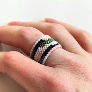 May include: A beaded ring with a colorful pattern. The ring features a repeating design of black, white, pink, and green beads. The ring is worn on a finger, showcasing the intricate beadwork and color combination.