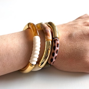 Peut inclure: Trois bracelets dorés avec des accents colorés sont portés au poignet. L'un des bracelets a un motif torsadé blanc, un autre un motif marbré marron et beige, et le troisième un imprimé léopard marron et rose.