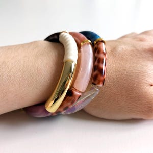 Könnte beinhalten: Eine Sammlung farbenfroher, klobiger Armbänder. Ein Armband hat ein weißes und goldenes Design, ein anderes einen rosa Stein und ein drittes ein Leopardenmuster. Die Armbänder werden am Handgelenk getragen.