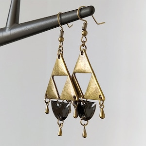 May include: Gold triangle earrings with black teardrop-shaped beads hanging from the bottom. The earrings have a geometric design.