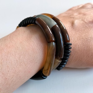 Può includere: Un set di braccialetti impilati nelle tonalità del marrone, ambra e grigio. I braccialetti presentano un mix di segmenti lisci e curvi ed elementi neri strutturati. I gioielli sono indossati su un polso, mostrando il design e le variazioni di colore.