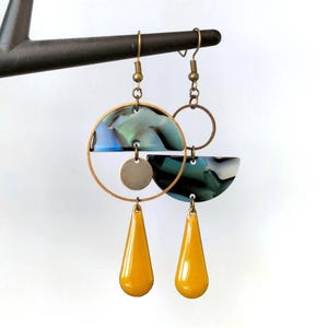May include: A pair of abstract earrings with a brass finish. Each earring features a half-circle and a teardrop shape in yellow, with a marbled blue and green design. A small brass disc and a brass ring complete the design.