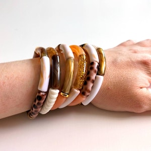 Könnte beinhalten: Eine Sammlung bunter Armreifen an einem Handgelenk. Die Armbänder weisen eine Vielzahl von Designs auf, darunter weiße, braune, orangefarbene, goldglitzernde und Leopardenmuster. Die Armreifen sind übereinander gestapelt und erzeugen einen auffälligen und modischen Look.