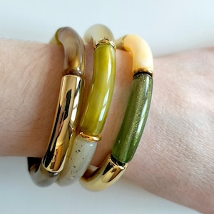 Op de afbeelding: Drie gouden armbandjes met groene en witte hars. De armbandjes zijn gestapeld op de pols van een persoon.