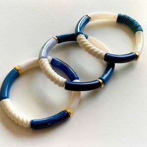May include: Three linked bracelets with a nautical theme. The bracelets feature a combination of white, navy blue, and light blue segments. Some sections have a twisted rope design, and all have gold-colored accents. The bracelets are on a white surface.
