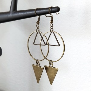 May include: A pair of gold-toned earrings with a circle and triangle design. The earrings are hanging from a black earring stand.