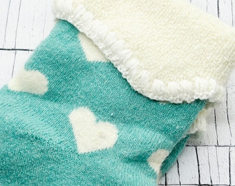 Puede incluir: Un calcetín de color verde azulado con un patrón repetitivo de corazones blancos. El calcetín tiene un puño grueso de color crema con un borde de ganchillo decorativo y texturizado en el dobladillo superior, contrastando con el tejido.