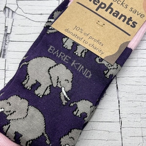 May include: A pair of socks with a purple background and a pattern of grey elephants. The socks have a pink cuff and a tag that reads "these socks save elephants." The tag also states that 10% of profits are donated to charity.