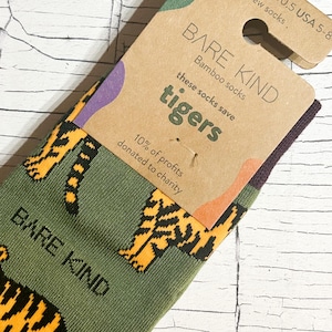 Könnte beinhalten: Grüne Socken mit einem Muster aus orangefarbenen Tigern. Die Socken sind mit einem braunen Papieranhänger verpackt, auf dem "BARE KIND Bamboo socks these socks save tigers 10% of profits donated to charity" steht.