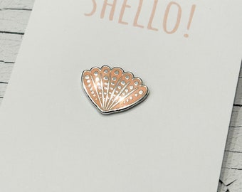 May include: A white card with the text "SHELLO!" in pink lettering. A small, peach-coloured enamel pin shaped like a seashell with white dots is attached to the card. The card also has the text "CORAL & MINT WITH LOVE" printed on the bottom.