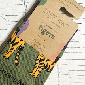 Könnte beinhalten: Grüne Socken mit einem Muster aus orangefarbenen Tigern. Die Socken sind mit einem braunen Kartonanhänger verpackt, auf dem "BARE KIND Bamboo socks these socks save tigers 10% of profits donated to charity" steht.