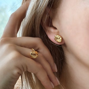 May include: Gold-colored jewelry set featuring a ring and stud earrings, each designed in the shape of a coffee bean. The ring is on a finger, and the earrings are on an ear. The jewelry has a textured surface and a shiny finish.