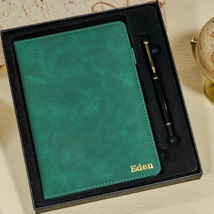 Puede incluir: Un juego de diario y bolígrafo de cuero verde en una caja de regalo negra. El diario tiene el nombre "Eden" en letras doradas. Un bolígrafo negro con detalles dorados descansa junto al diario. Un pequeño globo terráqueo es visible.
