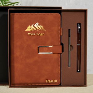 Op de afbeelding: Een bruin leren notitieboek met een gouden berglogo en de tekst "Your Logo". Het notitieboek zit in een geschenkdoos met een pen en een penvulling. Het notitieboek heeft een metalen sluiting en de naam "Paul" met een hart.